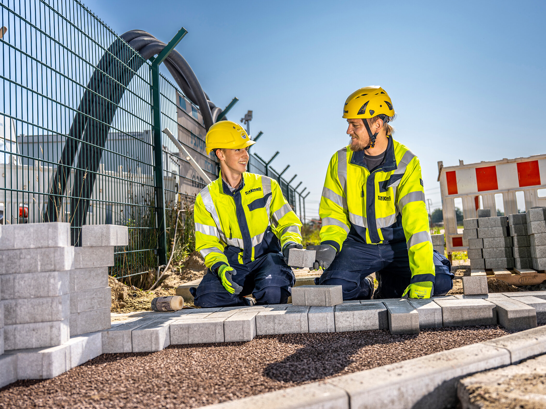 Zwei Auszubildende verlegen gemeinsam Pflastersteine auf einer Baustelle.