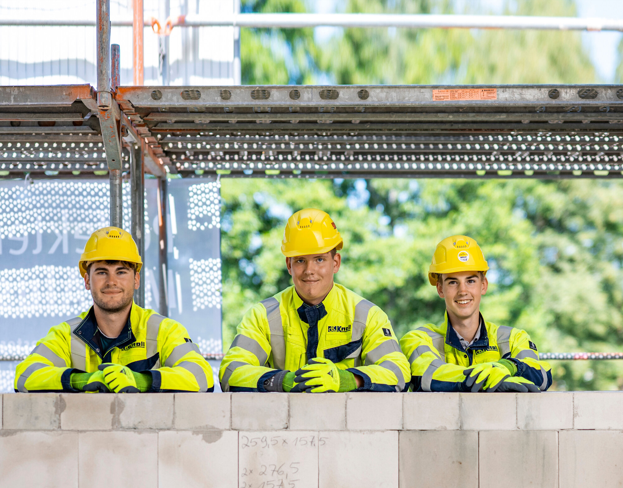 Drei Praktikanten oder Auszubildende in Arbeitskleidung stehen gemeinsam auf einer Baustelle und blicken in die Kamera.